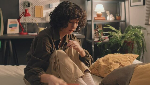 Young girl with curly short hair checking her phone, then putting it away, looking depressed and disappointed, touching her neck and face, sitting on unmade bed, desk, lamps and plant in background