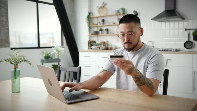 Man Paying Online Using Laptop. Asian 30s man using bank credit card for online shopping and purchase sitting alone in apartments. Close Up.