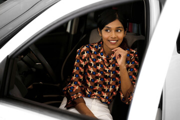 Attractive smiling Indian woman sitting inside new car looking away in auto salon. Transportation...