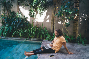 Concentrated man on vacation near pool working on laptop