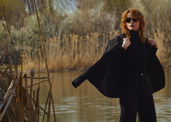 a stylish woman in a black suit and sunglasses walks in the park near the lake
