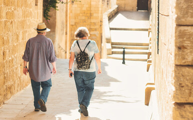 An unrecognizable mature white Caucasian couple of tourists walking through a typical Mediterranean village - wearing backpack and straw panama hat