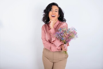 Optimistic young woman holds big bouquet of nice flowers over white background keeps hands partly crossed and hand under chin, looks at camera with pleasure.