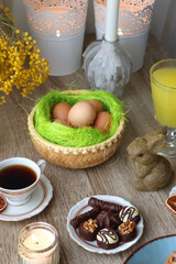 Easter eggs in the basket, bowl of cookies, chocolate pralines, Easter bunny figurine, cups of tea, glasses of juice, flowers and lit candles on the table. Selective focus.