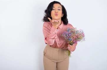young woman holds big bouquet of nice flowers over white background looking at the camera blowing a kiss with hand on air being lovely and sexy. Love expression.