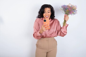young woman holds big bouquet of nice flowers over white background holding in hands cell and rising his fist up being excited after reading good news.
