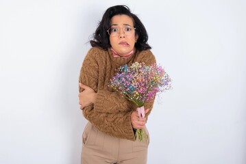 young woman holds big bouquet of nice flowers over white background shaking and freezing for winter cold with sad and shock expression on face.