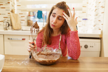 Blonde middle-aged woman with a bowl in front of her in the kitchen is having a problem with her cake. Woman in kitchen with dough beater looking lost in thought.