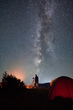 Professional photo shoot of Milky Way in mountains. Male photographer taking photos of starry night in mountains. Back view of man with camera between next to his tent.