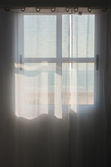 Window to the Sea. Transparent curtain with the shadow of the silhouette of the window and sunlight. View through the curtains to the sea and beach. Concept interior and vacation.