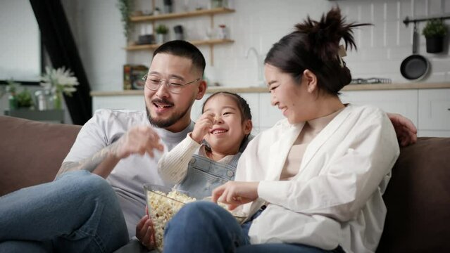 Family Watching TV Eating Popcorn. Korean family with one kid having fun sitting on couch at home. Happy mum and dad laughing with daughter.