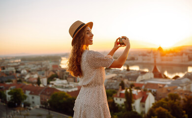 Fototapeta premium Tourist woman shoots a city landscape at dawn on a smartphone. Travel in Europe. A young woman enjoys the sunrise. The concept of travel, tourism, vacation and freedom.