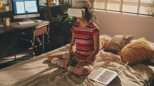 Full shot of short-haired young girl in white VR headset, wearing bright T-shirt and shorts sitting on her bed in morning, her hand on laptop, moving her head. Sunlit bedroom