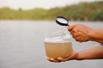 Closeup hands hold megnifying glass to inspect water in jar that contain sample water from the...