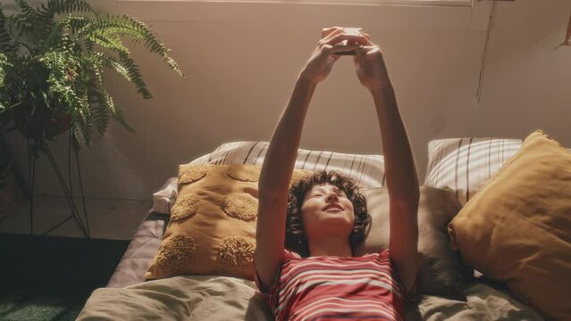 Tight medium shot of short-haired curly girl in bright stripped T-shirt lying in unmade bed, making video or selfie with her smartphone, green plant beside her. Sunlit cozy bedroom, morning