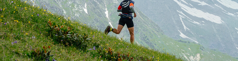 male runner run down mountain trail, skyrunning race ultramarathon, green meadows and snow-capped...