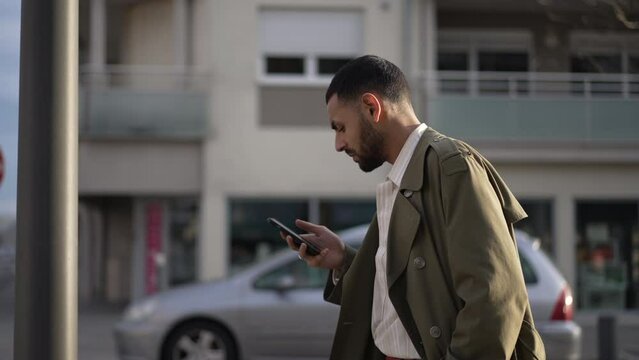 One young Arab man holding phone while walking in street with serious expression. A Middle Eastern man wearing jacket reading text message