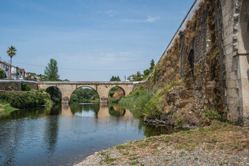 Fototapeta na wymiar Wall of ancient stone bridge with herbs and medieval bridge of Coja in the background reflected in the river Alva, Arganil PORTUGAL