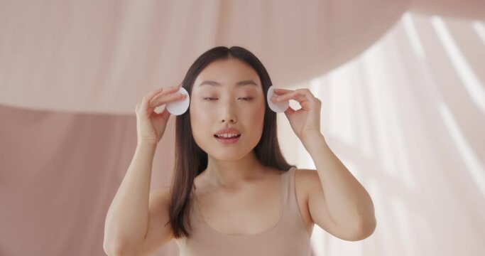 Beautiful Asian woman cleaning face skin. Pretty young female using cotton pad, smoothing skin and removing makeup with micelar water or lotion. Puring, washing, wiping. Skin care, beauty concept.