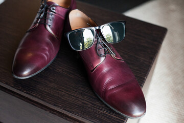 The groom's burgundy men's shoes and glasses stand on a textured dark wooden surface in the room. Image for your creative design or illustrations.