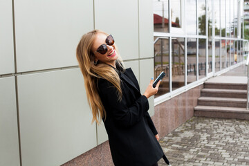 A beautiful young girl with blond hair in sunglasses and a black jacket stands on a city street with a phone in her hands and laughs.