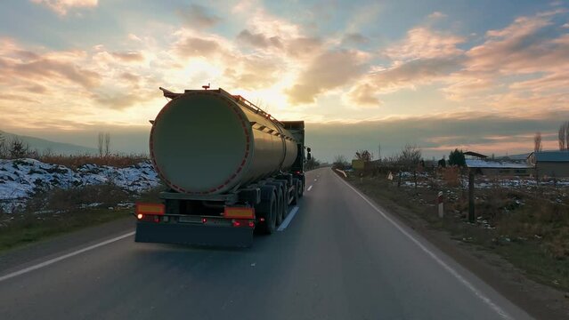 A big metal fuel tanker truck shipping fuel on the countryside road in motion against a blue sky with a sunset