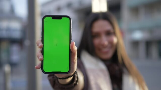 One young woman showing content online to camera while holding smartphone with Chroma Key Greenscreen standing in city street with one hand