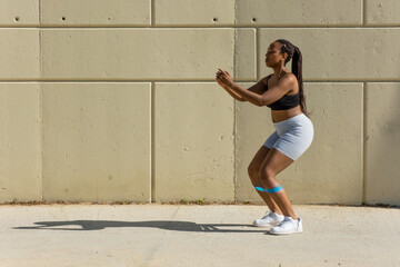 performing sport outdoors, young black girl performing squats with elastic tape on legs in...