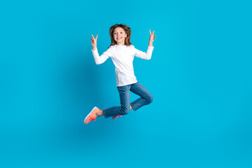 Fototapeta na wymiar Full length photo of cheerful adorable schoolgirl dressed white shirt jeans flying showing v-sign isolated on blue color background
