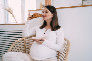 Happy pregnant woman sniffing fresh croissant in morning