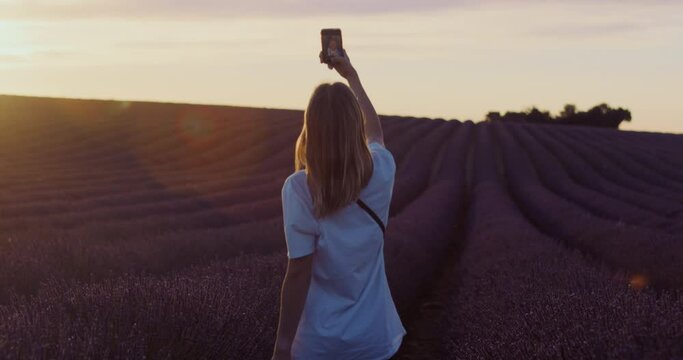 Role model or blogger takes selfies on phone in lavender fields at sunrise or sunset. Handheld shot. Fresh breeze in hair. Outdoor adventure. Freedom lifestyle. Influencer at famous spot. 