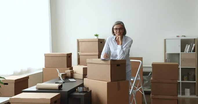 Confident mature woman retail seller, individual entrepreneur smile looks at camera, pose in dropshipping storage, boxes prepared for shipment nearby. Delivery services, small business owner portrait