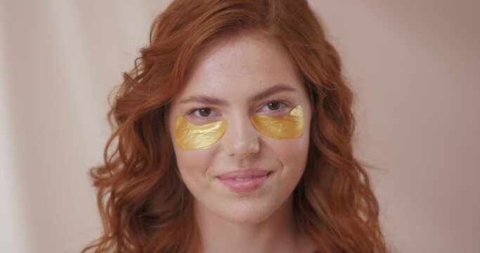 Adorable curly woman glancing straight on camera. Happy woman smiling and using gold eye patches with collagen, peptide, ceramide and hyaluronic acid, derma oil. Dermatology, healthy skin concept.