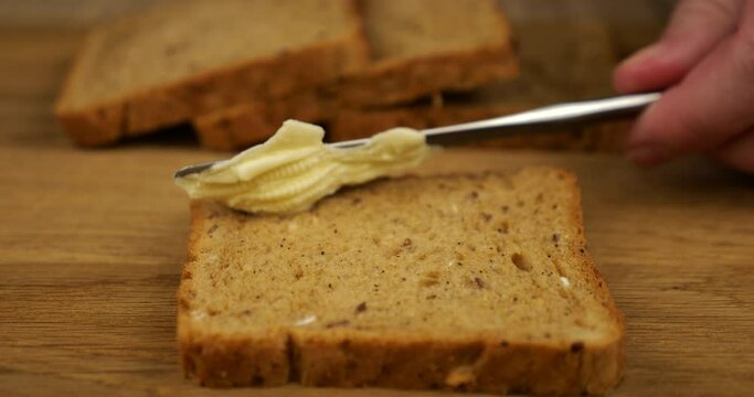 Butter is spread with a knife on a piece of multigrain bread on a wooden cutting board.