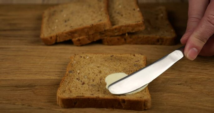 Butter is spread with a knife on a piece of multigrain bread on a wooden cutting board.