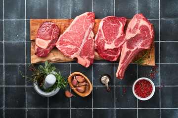 Raw prime steaks. Variety of fresh black angus prime meat steaks T-bone, New York, Ribeye, Striploin, Tomahawk on black old tiles stone background. Set of various classic steaks. Top view copy space.