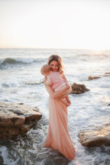 Fototapeta na wymiar Full length portrait of a beautiful fairytale mother huging her infante daughter in the summer backdrop of waves at sunset.Woman in a long delicate pink dress, a child up to a year old in pink clothes