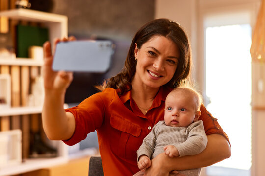 A smiling mother takes selfies with a phone with her adorable baby girl at home.