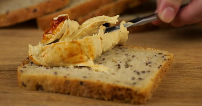 Hummus is spread with a knife on a piece of multigrain bread on a wooden cutting board. Food for vegans.