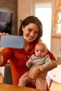 A smiling mother holding a baby girl and taking a selfie photo at home.