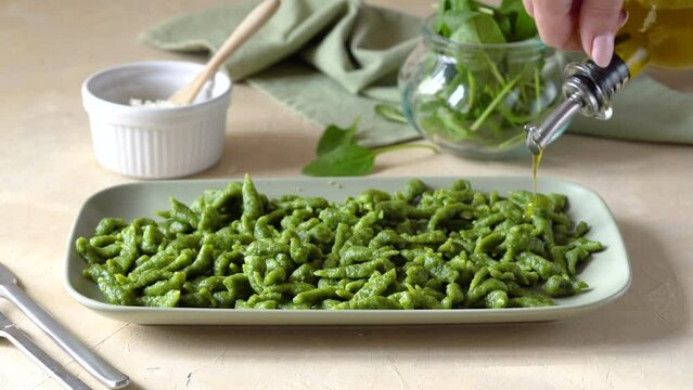 Spinach spatzle are the typical Tyrolean green dumplings or gnocchi on green plate with grated cheese and olive oil. Woman's hand is pouring olive oil and cheese to the dish
