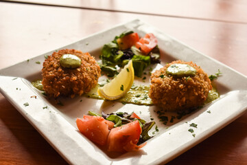 crab cakes on a square plate with green sauce, lemon, greens, and tomato