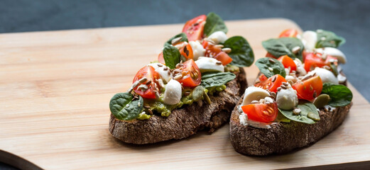 Bruschetta (sandwiches) with cherry tomatoes, mozzarella cheese and herbs on a cutting board on a dark background. Traditional Italian snack.