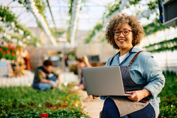 Happy woman using laptop while working in greenhouse and looking at camera.
