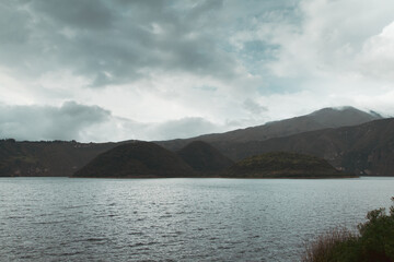 large plume of rain clouds in the distance as a mountain rage is seen from the shores of a crater lake of an inactive volcano, the sun shines on the waters surface through the clouds
