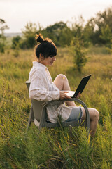 a young woman wearing a shirt works on a laptop while sitting on a chair in the middle of nature