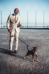 Vertical shot of a bald black man with a beard walking his dog on Lisbon's cobblestone pavement. Holds the leash in one hand and his sunglasses in the other, wearing a stylish suit and a colorful tie