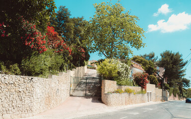 A bright, sunny day in Estoril, an entrance to a villa from across the street, with a sandstone wall, a green gate, and vibrant blooming plants and trees, all set against the blue sky