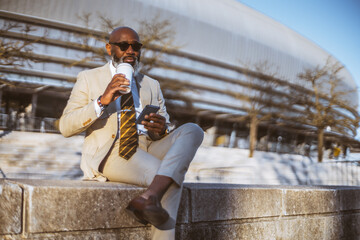 A bald man with a full white-bearded face holding a phone and coffee, dressed in a cream suit with...