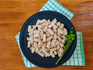 Raw tempeh cut placed on black plate with wooden background. Ready to cook

